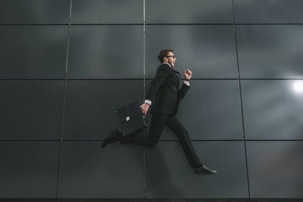 excited stylish businessman in suit and sunglasses jumping in front of black wall